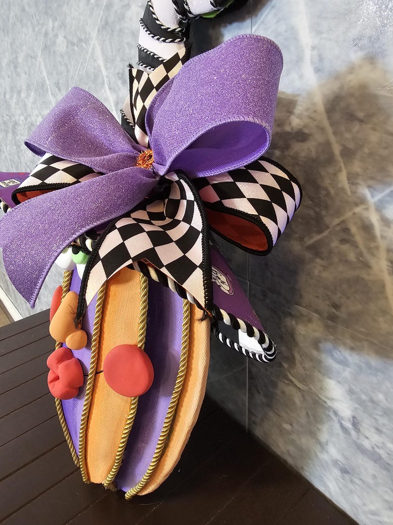 Pu&ograve; includere: Una decorazione da parete con un grande fiocco viola glitterato sovrapposto a un nastro a scacchi bianchi e neri. La base &egrave; una forma di zucca viola e arancione, arricchita da un bordo in corda dorata e piccole forme in argilla rosse.