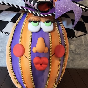 Pu&ograve; includere: Una zucca decorativa con segmenti verticali alternati viola e arancioni, rifiniti con cordoncino dorato. Il volto presenta occhi verdi con lunghe ciglia nere, un naso color pesca, guance e labbra rosse, sormontato da un grande fiocco a scacchi viola e nero.