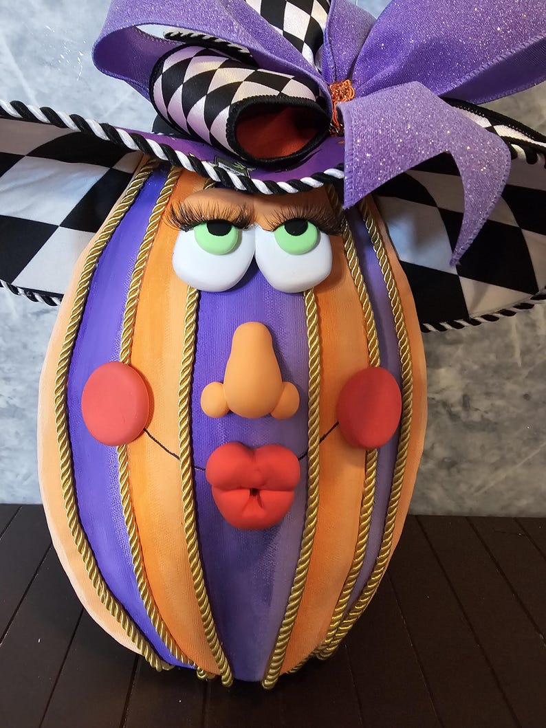 Pu&ograve; includere: Una zucca decorativa con segmenti verticali alternati viola e arancioni, rifiniti con cordoncino dorato. Il volto presenta occhi verdi con lunghe ciglia nere, un naso color pesca, guance e labbra rosse, sormontato da un grande fiocco a scacchi viola e nero.