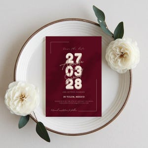 Op de afbeelding: Een bordeauxrode huwelijksuitnodiging met de tekst "Save the Date" en de datum "27 03 28". De uitnodiging bevat de tekst "Are getting married in Tulum, Mexico". De uitnodiging ligt op een wit bord met witte bloemen.