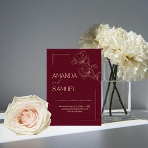 Op de afbeelding: Een bordeauxrode huwelijksuitnodiging met gouden tekst en bloemenlijntekeningen. De uitnodiging luidt "AMANDA en SAMUEL" en bevat huwelijksdetails. Een witte roos en een boeket witte bloemen in een glazen vaas staan ook op de afbeelding.