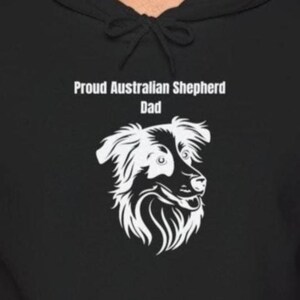 Peut inclure: Sweat à capuche noir avec un graphisme blanc d'un chien berger australien et le texte "Proud Australian Shepherd Dad". L'illustration du chien est détaillée avec un accent sur le visage et la fourrure.