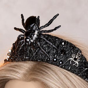 May include: A black tiara featuring a prominent central spider ornament with a faceted black stone body, surrounded by smaller silver-toned spider accents and intricate black textured webbing patterns embellished with tiny clear crystals.