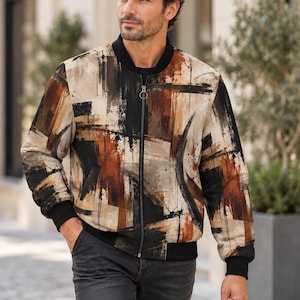 May include: A bomber jacket featuring an abstract brushstroke pattern in shades of black, brown, and beige. The jacket has a black ribbed collar, black ribbed cuffs, and a central silver-toned metal zipper closure with a circular pull tab.