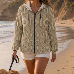 May include: A light beige zip-up hooded jacket featuring a repeating pattern of dog bones and paw prints in a darker shade. The jacket has long sleeves with elastic cuffs, a black front zipper, and a white inner lining visible at the collar area.