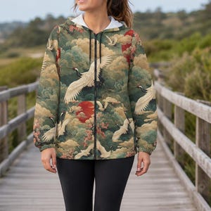 May include: A zip-up hooded jacket featuring a repeating pattern of white cranes in flight, red suns, and green clouds. The jacket has long sleeves, a front zipper closure, and a white inner lining visible at the neck. It is paired with black leggings.