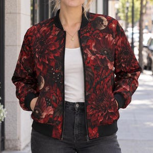 May include: A bomber jacket featuring a dark floral pattern with deep red and black flowers, interspersed with small moon and star motifs. The jacket has a black ribbed collar, black cuffs, a black hem, and a central black zipper closure.