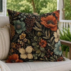 May include: A square throw pillow featuring a dark black background with a dense botanical print of orange poppies, muted teal flowers, yellow blossoms, and green foliage. The pillow is positioned next to a plain, light beige textured throw pillow.