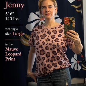 May include: A short-sleeved crew neck t-shirt featuring a mauve leopard print pattern with dark brown and black spots. The shirt is shown in a size Large, as indicated by the text overlay, and is paired with blue denim jeans.