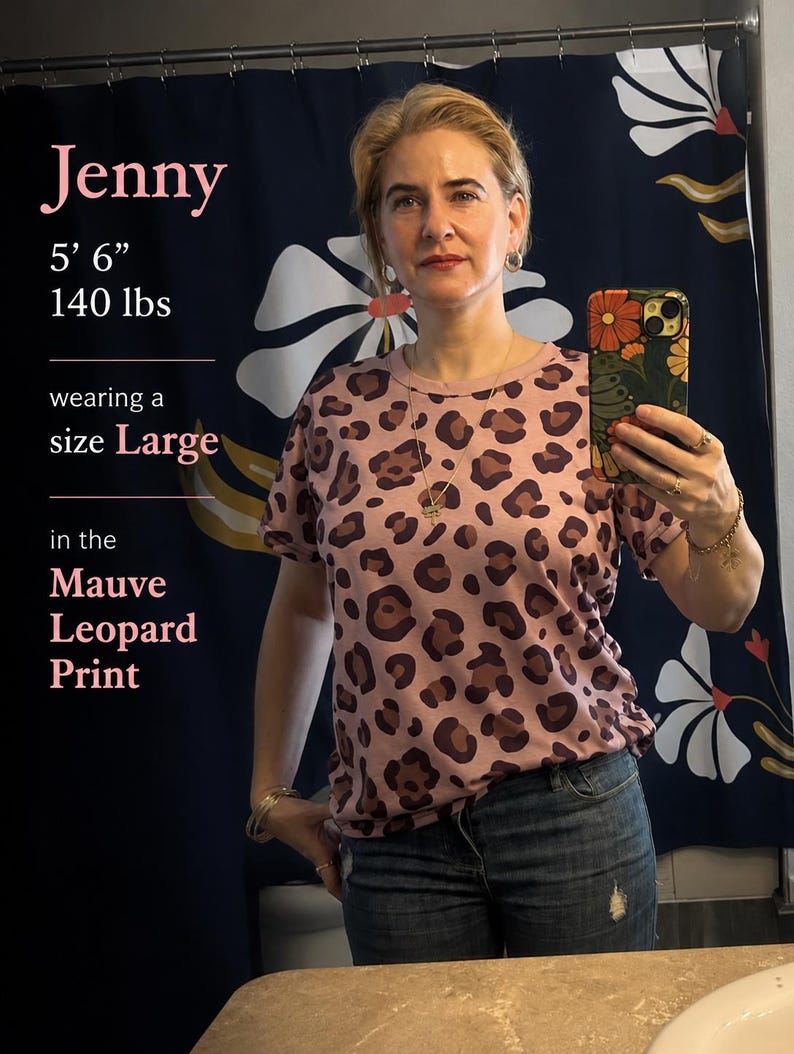 May include: A short-sleeved crew neck t-shirt featuring a mauve leopard print pattern with dark brown and black spots. The shirt is shown in a size Large, as indicated by the text overlay, and is paired with blue denim jeans.