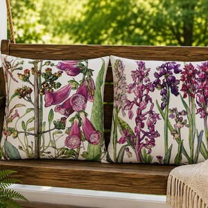 May include: Two square throw pillows featuring botanical illustrations on a white background. The left pillow displays pink foxglove flowers, while the right pillow showcases purple orchid-like blooms with green stems and leaves on both covers.