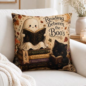 May include: A square throw pillow featuring a cream-coloured background with a ghost reading a book atop a stack of four books, next to a black kitten reading a small book. The text reads Reading Between the Boo's, with autumn leaves and mushrooms.