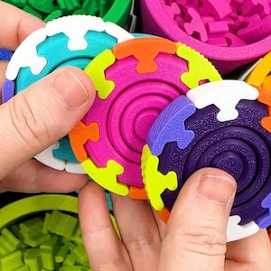 May include: Three circular, gear-shaped fidget toys featuring interlocking puzzle pieces in vibrant colors including teal, magenta, and purple. Each toy has a textured, concentric ring pattern on the center surface and is surrounded by smaller, contrasting colored plastic segments in white, lime green, and orange.