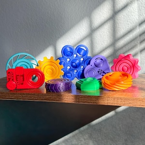May include: A collection of colorful 3D-printed fidget toys in various shapes, including gears, wheels, and abstract designs. The toys are in shades of red, yellow, blue, purple, green, orange, and pink, arranged on a wooden surface.