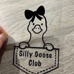 May include: Black graphic of a goose wearing a bow, peeking out of a pocket that reads "Silly Goose Club". The design is on a clear, transparent material, suitable for application on various surfaces. The goose has a cute, cartoonish appearance.