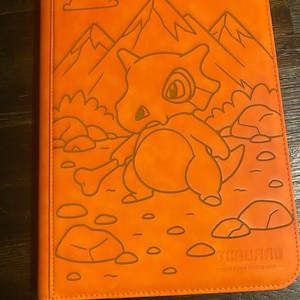 May include: Orange trading card binder with a Cubone character design, mountains, and rocks. The binder has a debossed design and the text "TCGUARD - guard your trading cards".