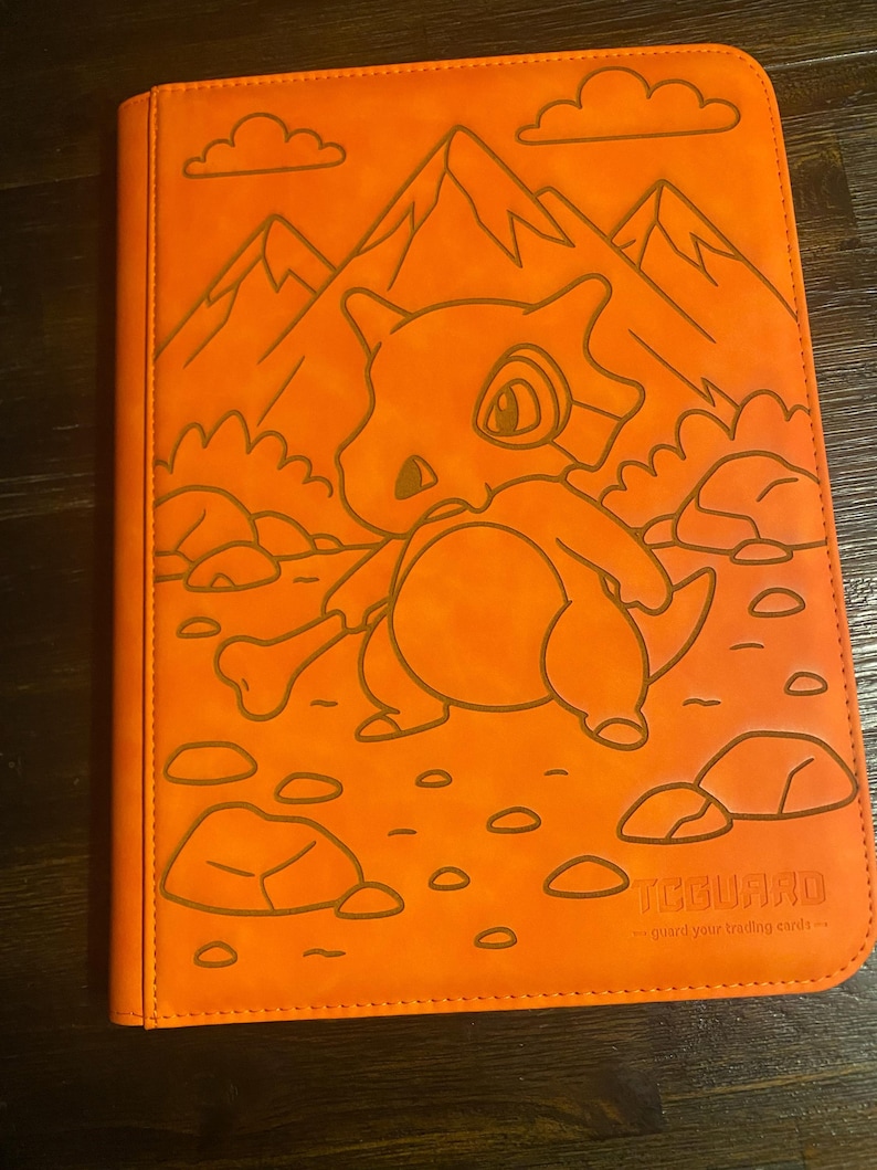 May include: Orange trading card binder with a Cubone character design, mountains, and rocks. The binder has a debossed design and the text "TCGUARD - guard your trading cards".