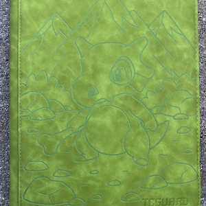 May include: Green trading card binder featuring embossed artwork of a cartoon character, mountains, clouds, and rocks. The binder has the text "TCGARD" and "- guard your trading cards -" at the bottom.
