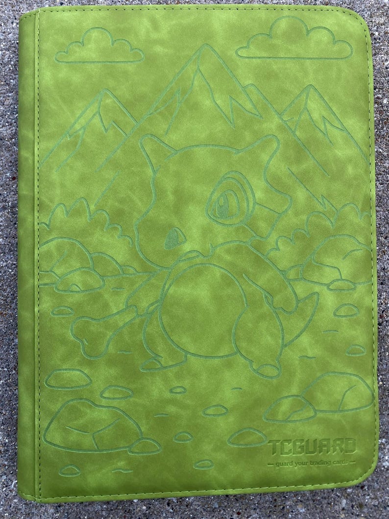 May include: Green trading card binder featuring embossed artwork of a cartoon character, mountains, clouds, and rocks. The binder has the text "TCGARD" and "- guard your trading cards -" at the bottom.