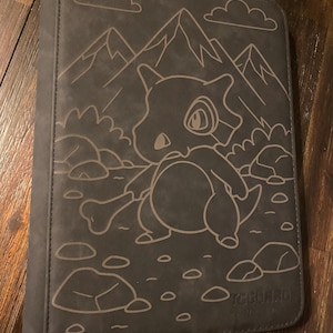 May include: A dark grey card binder with a detailed outline of a cartoon Cubone character holding a bone, set against a mountain and cloud backdrop. The binder is designed to hold trading cards.