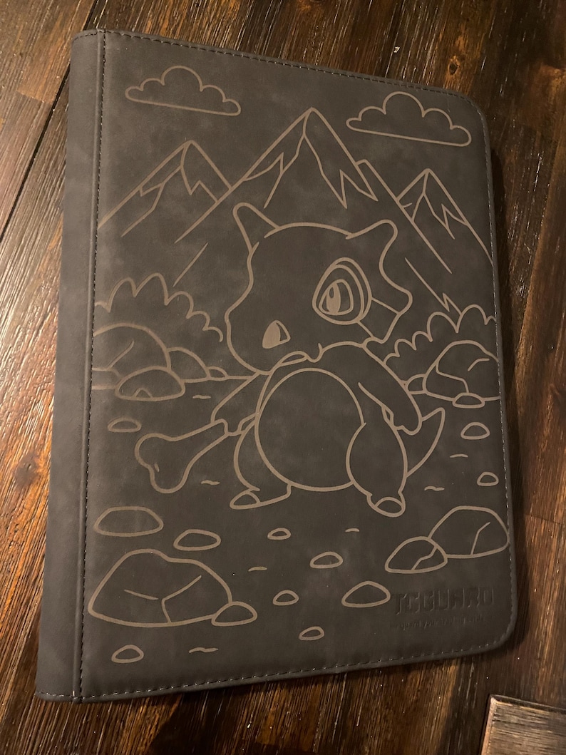 May include: A dark grey card binder with a detailed outline of a cartoon Cubone character holding a bone, set against a mountain and cloud backdrop. The binder is designed to hold trading cards.