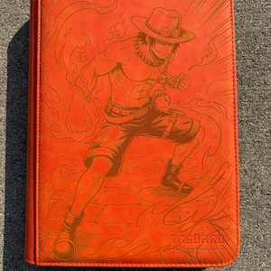 May include: A bright orange trading card binder featuring an embossed illustration of a character wearing a hat and shorts surrounded by flames. The cover includes the text TCGUARD and the phrase guard your trading cards in the bottom right corner.