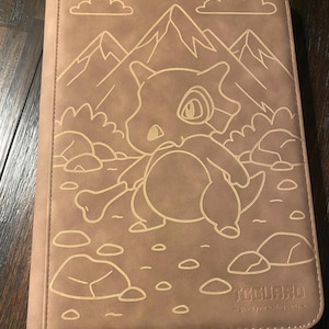May include: A tan trading card binder with a Cubone character design, mountains, and clouds. The binder has a stitched border and the text "TCGUARD - guard your trading cards."