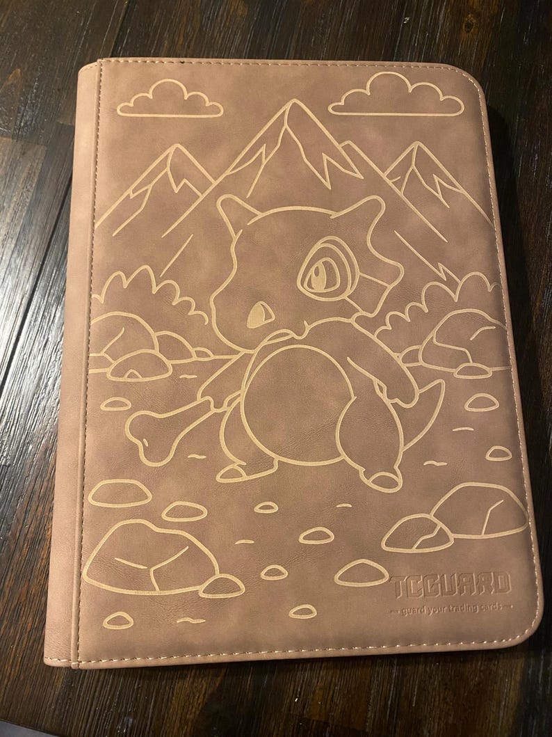 May include: A tan trading card binder with a Cubone character design, mountains, and clouds. The binder has a stitched border and the text "TCGUARD - guard your trading cards."