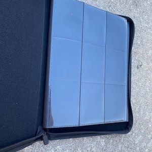 May include: Black zipped binder with a grey card holder insert. The card holder has multiple clear pockets for storing cards. The binder is open, revealing the interior and the zip is partially closed.
