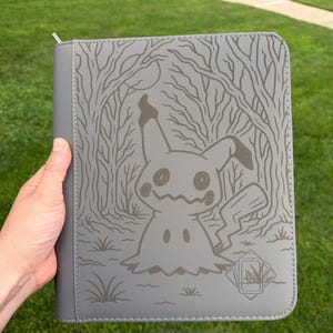 May include: A gray zippered binder featuring a laser-engraved illustration of the character Mimikyu standing in a forest. The design includes a full moon, bare trees, and a small emblem in the bottom right corner depicting a card inside a frame.