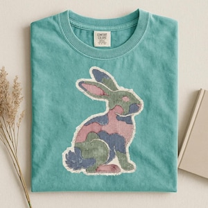 May include: A turquoise Comfort Colors t-shirt featuring a patchwork rabbit design. The rabbit is composed of pink, green, and blue fabric pieces with a frayed edge. The shirt has a crew neck and a small tag on the upper chest.