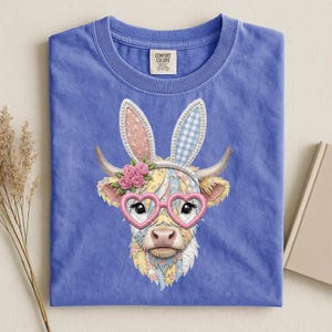 Puede incluir: Camiseta azul Comfort Colors con un diseño caprichoso de vaca. La vaca lleva gafas rosas en forma de corazón, orejas de conejo y una corona floral. El diseño incorpora colores pastel y estampados florales.