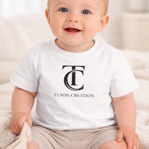 May include: A smiling baby wearing a white t-shirt with the black text "TUSON CREATION". The shirt features a stylized "TC" logo. The baby is also wearing beige shorts and white socks.