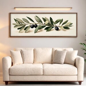 May include: A rectangular wall art piece featuring a horizontal olive branch with green leaves and three dark purple olives on a cream background, framed in wood. Below it sits a cream-colored sofa with a subtle floral pattern and five throw pillows.