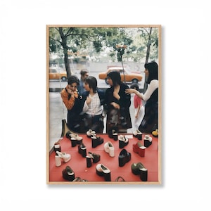 May include: A framed vintage photograph of a shoe store display. The display table is red and features various platform shoes in black, white, and red. Several people are looking at the shoes through the store window.