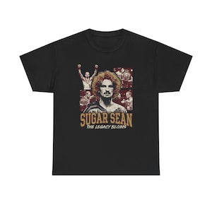 May include: Black t-shirt featuring a portrait of a man with curly hair, surrounded by smaller images. The text "SUGAR SEAN" is above "THE LEGACY BEGINS" in gold lettering. A sports-themed graphic tee.