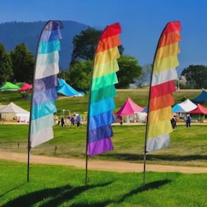 May include: Three rainbow-coloured feather flags stand on a grassy field. The flags are arranged in a row, with a variety of colours including blue, green, yellow, and red. In the background, there are tents and trees under a blue sky.