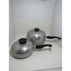 Op de afbeelding: Twee ronde aluminium steelpannen met bijpassende deksels en zwarte handgrepen. Elke handgreep heeft een kleine metalen lus aan het uiteinde om op te hangen. De pannen hebben een geborstelde metalen afwerking en cirkelvormige ribbels op de deksels.