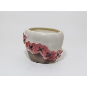 May include: A ceramic planter with a rounded shape, featuring a glossy cream-coloured upper section and a smooth brown base. A band of raised, three-dimensional pink flowers with dark centres wraps around the middle of the exterior surface.
