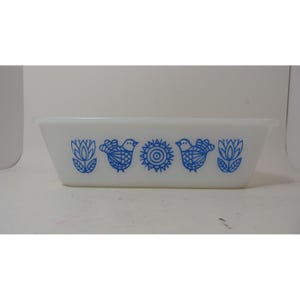 Könnte beinhalten: Eine rechteckige Backform aus weißem Glas mit einem wiederkehrenden blauen Muster aus stilisierten Blumen, Vögeln und einem zentralen Sonnenmotiv. Die Form hat eine glatte, opake Oberfläche, einen ausgestellten Rand und eine schlichte Form.