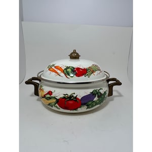 Könnte beinhalten: Weißer Emaille-Topf mit Deckel, mit einem farbenfrohen Gemüse-Design. Der Topf hat bronzefarbene Griffe und einen passenden Knauf auf dem Deckel. Gemüse umfasst Karotten, Tomaten, Paprika und Spargel. Ein dekorativer Küchenartikel.