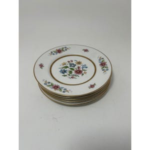 Puede incluir: Una pila de platos de cerámica blanca con un ramo floral central de flores rojas, azules y amarillas. Cada plato tiene un círculo interior y un borde dorados, con pequeños motivos florales dispersos por el borde blanco exterior.