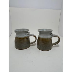 Peut inclure: Deux mugs en céramique faits à la main. Les mugs ont une base arrondie, un corps cylindrique et une anse incurvée. Le haut des mugs est de couleur gris clair, passant au marron puis à un gris plus foncé à la base. Les mugs sont sur une surface blanche.
