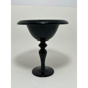 May include: A black glass pedestal bowl with a wide, shallow rim. The bowl has a rounded body and sits atop a slender stem with a bulbous, decorative design. The base is a circular foot. The bowl is likely used for serving.