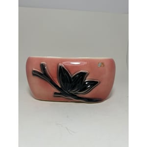 May include: A rectangular, pink ceramic planter with a black floral design. The planter has a curved shape and a glossy finish. The black floral design is a stylized depiction of a flower and leaves.