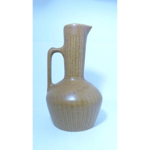 May include: A tan ceramic pitcher with a speckled finish, featuring a rounded base and a tall, slender neck. The body and neck are decorated with vertical ribbed detailing, and a single handle is attached to the side of the neck and shoulder.