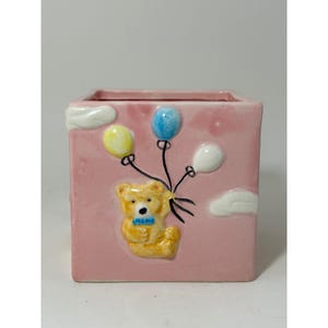 Könnte beinhalten: Ein rosa quadratischer Keramiktopf mit einem erhabenen Design eines Teddybären, der Luftballons hält. Die Ballons sind gelb, blau und weiß. Weiße Wolken befinden sich an den Seiten. Der Teddybär hat eine blaue Fliege.