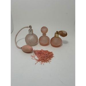 May include: Three antique perfume bottles in varying shades of pink. The bottles feature a ribbed design and are finished with silver and gold detailing. One bottle has a bulb atomizer and a pink tassel.