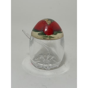 May include: A clear glass condiment jar with a rounded base and a domed lid featuring a painted red strawberry design on a cream background. The jar includes a small, clear glass spoon resting inside, and the base has a moulded floral pattern.