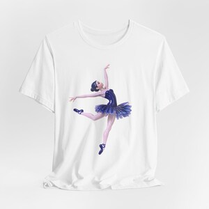 Peut inclure: Un t-shirt blanc à manches courtes avec une impression graphique représentant une ballerine vêtue d'un justaucorps bleu foncé et d'un tutu à volants. La danseuse est représentée en plein saut, les bras étendus et portant des chaussons bleus.
